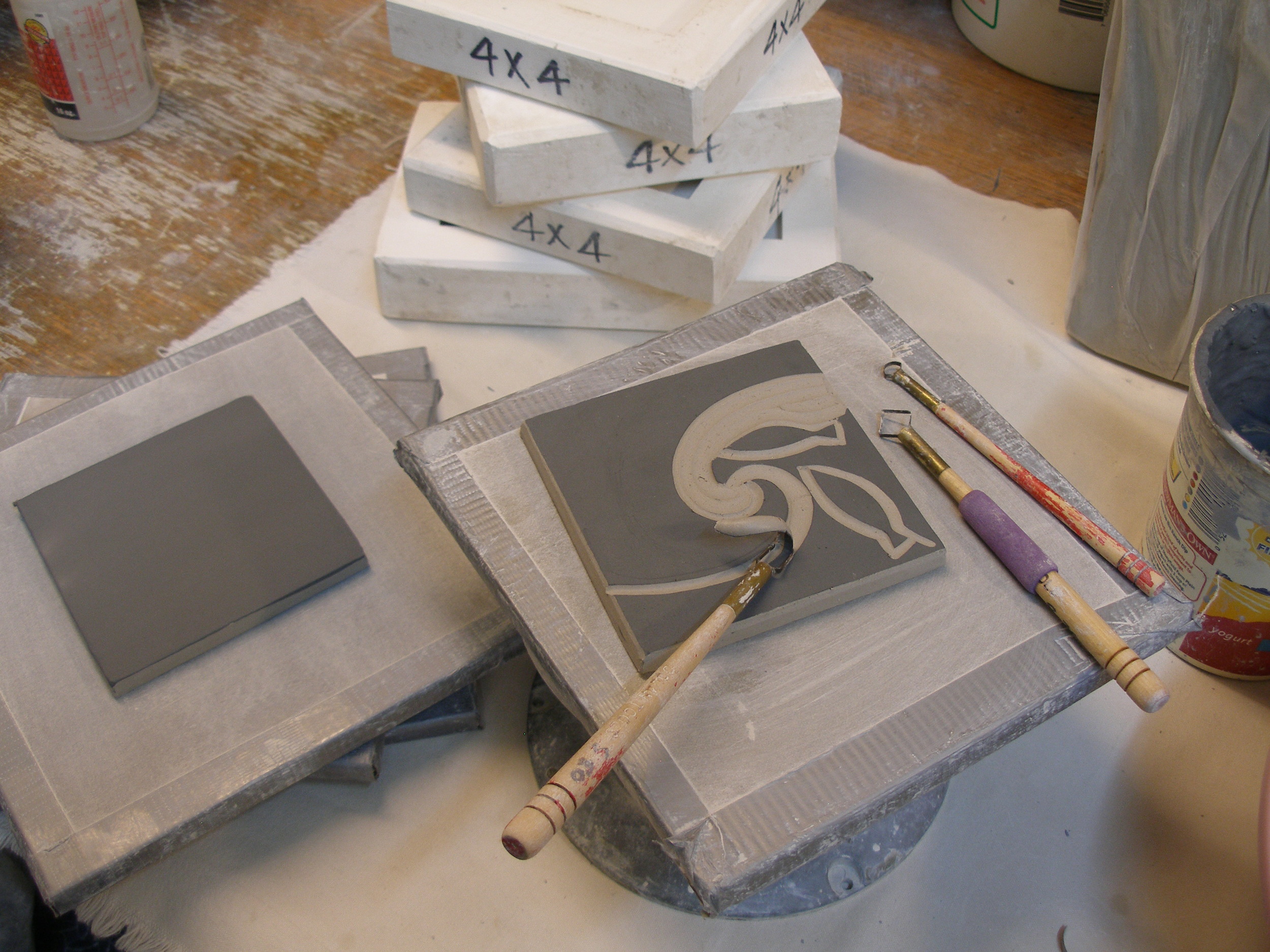 The blank tile is brushed with a layer of black colored clay, or slip, and once the slip has dried, stacked between boards to firm up overnight. When the tile has reached the leather-hard stage, still pliable, but easily retaining it's shape, it is ready to carve. The design is done free-hand, using a variety of looped cutting tools to remove the background, leaving the design. The carved tile is then stacked again between boards and left to dry for at least a week before it is dry enough for the bisque firing.