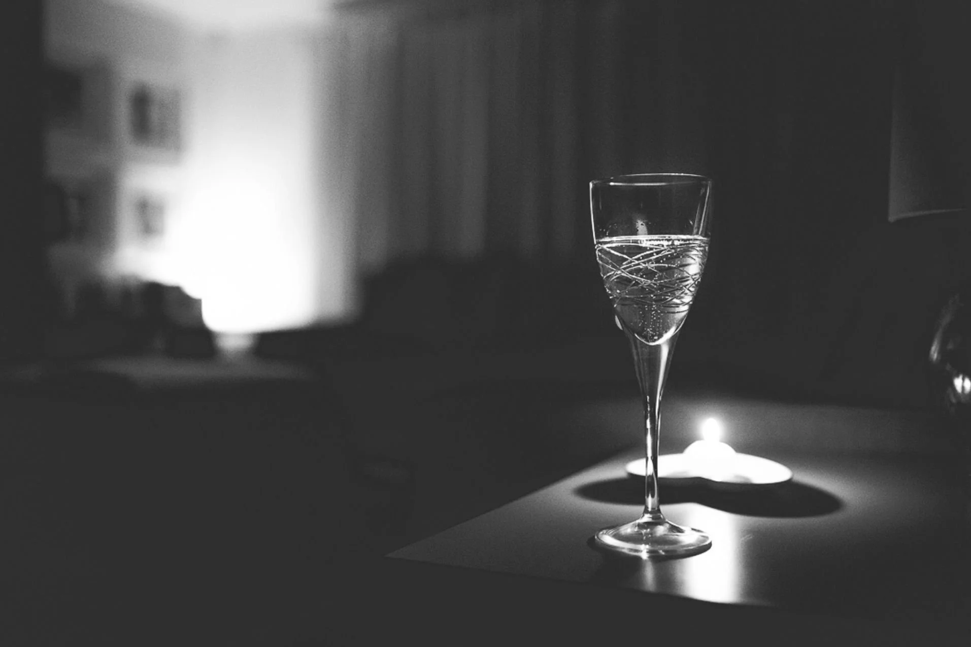Wine glass illuminated by the warm glow of a candle, with a small lamp providing additional light in the background.