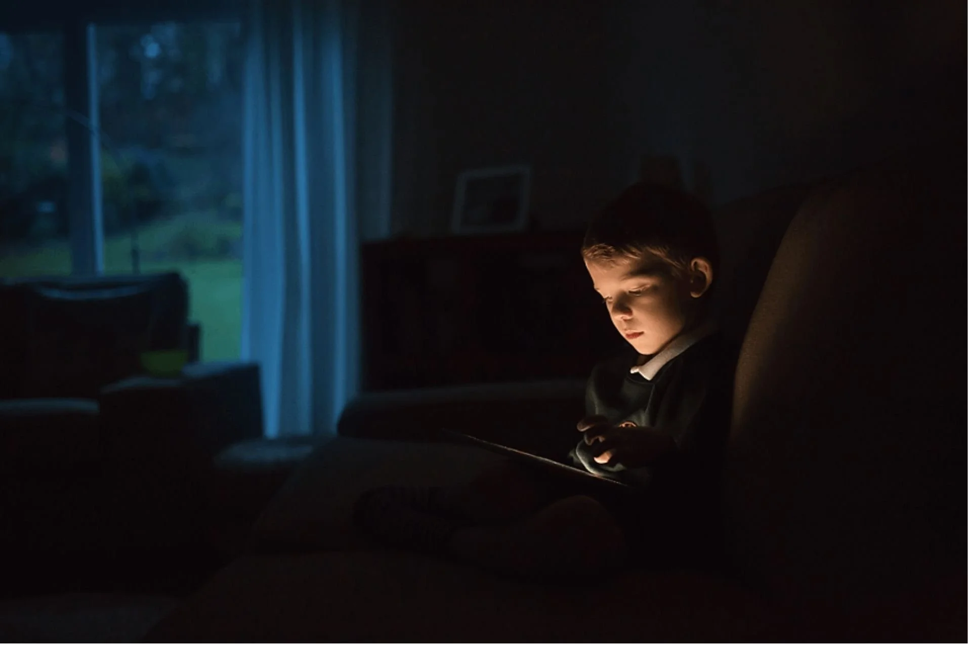 Young boy sitting on a sofa in a dark living room, his face illuminated by the warm glow of an iPad, with blue evening light coming through the window.