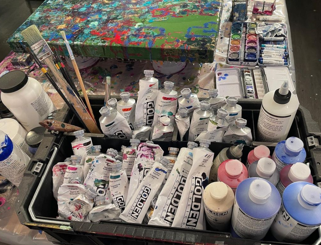 A table with art supplies including tubes of Golden acrylic paint, paint bottles, brushes, and palettes, with a splattered abstract painting on a canvas.