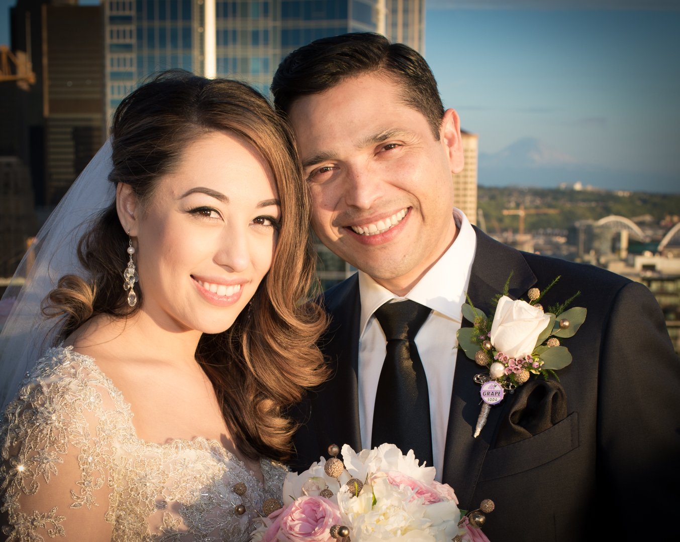 A newlywed couple smiling for a photo on a rooftop, dressed in wedding attire, with city buildings and a mountain in the background.