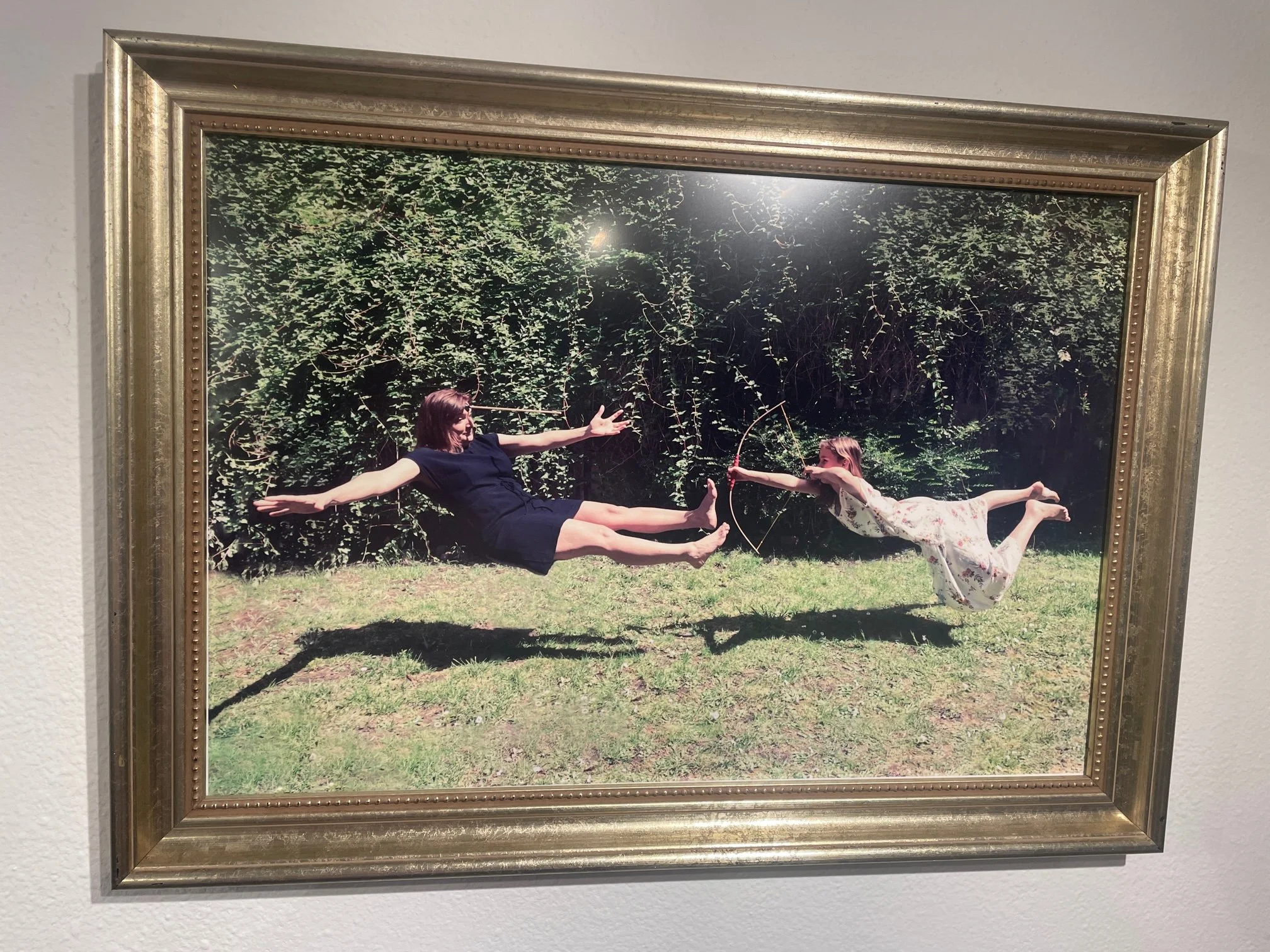 A framed photo of two women levitating outdoors, one holding a bow and arrow, with a leafy background and grass ground.