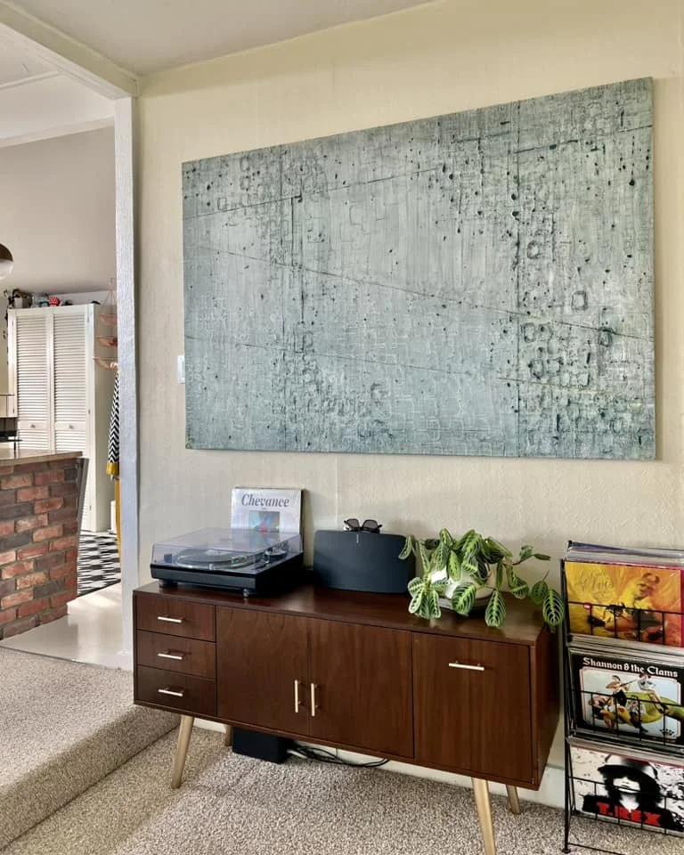 Living room with a large textured artwork on the wall above a wooden sideboard. The sideboard holds a turntable, a plant, and a magazine rack with vinyl records.