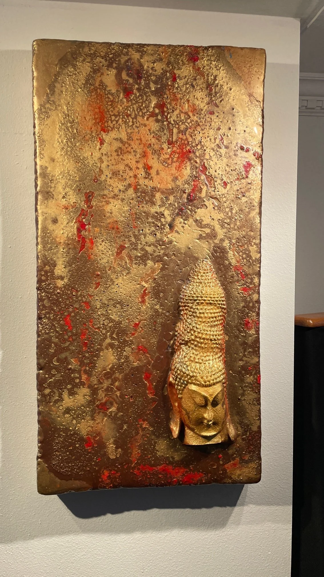 A textured metallic wall art piece with a reddish-brown color and abstract patterns, featuring a small carved wooden African mask sculpture attached near the bottom right.