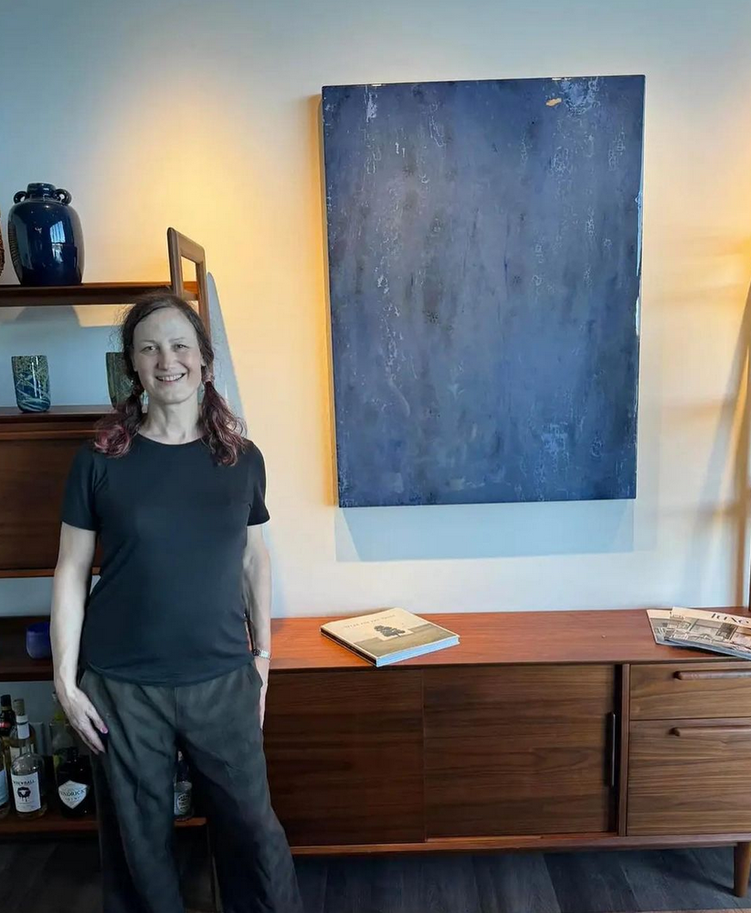 A woman with shoulder-length hair, wearing a black t-shirt and patterned pants, standing and smiling in a modern living space. Behind her is a wooden sideboard with books and magazines on top, and decorative vases on a shelf. A large blue abstract painting hangs on the white wall.