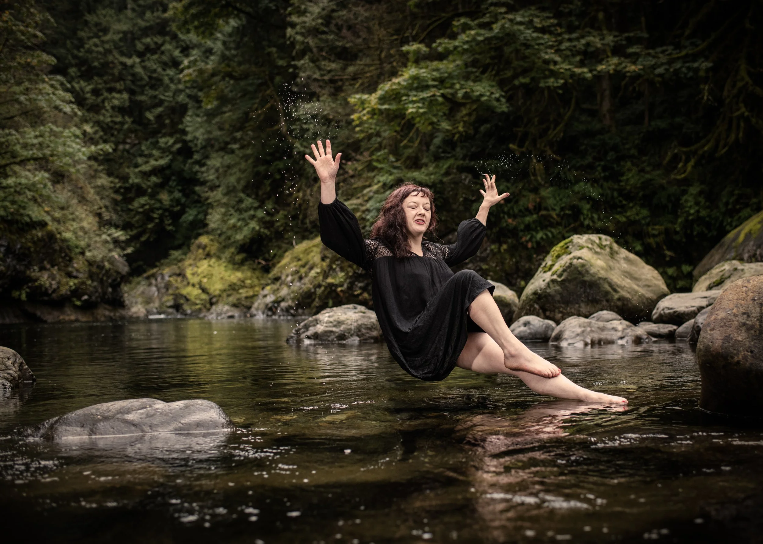 A woman in a black dress sitting on a rock in a river with her arms raised and water splashing around her, surrounded by greenery and moss-covered rocks.