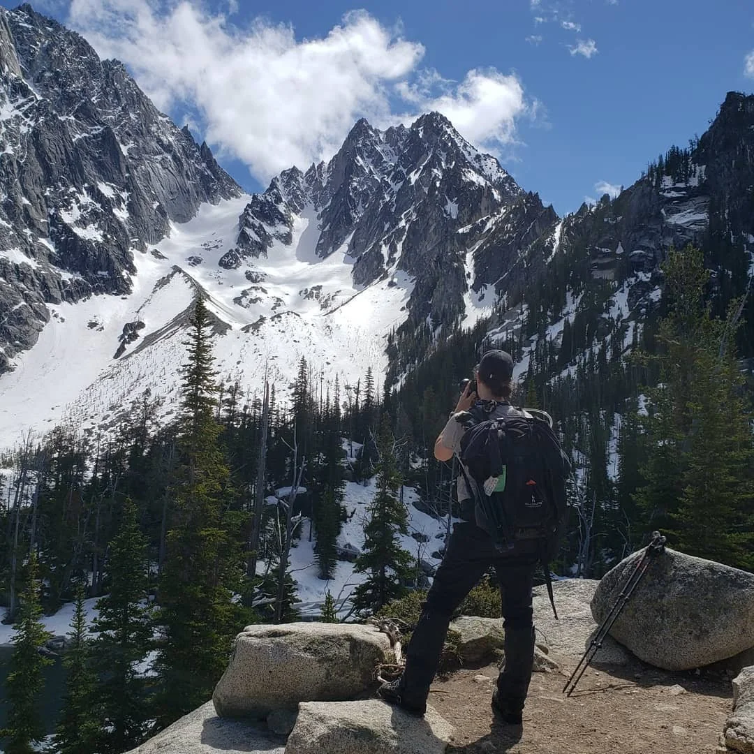 A hiker standing on a rocky ledge taking a photo of snow-covered mountain peaks in a scenic alpine landscape.