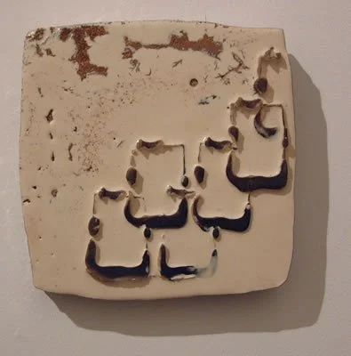 A white tile with dark, smudged Arabic calligraphy on it, mounted on a wall.