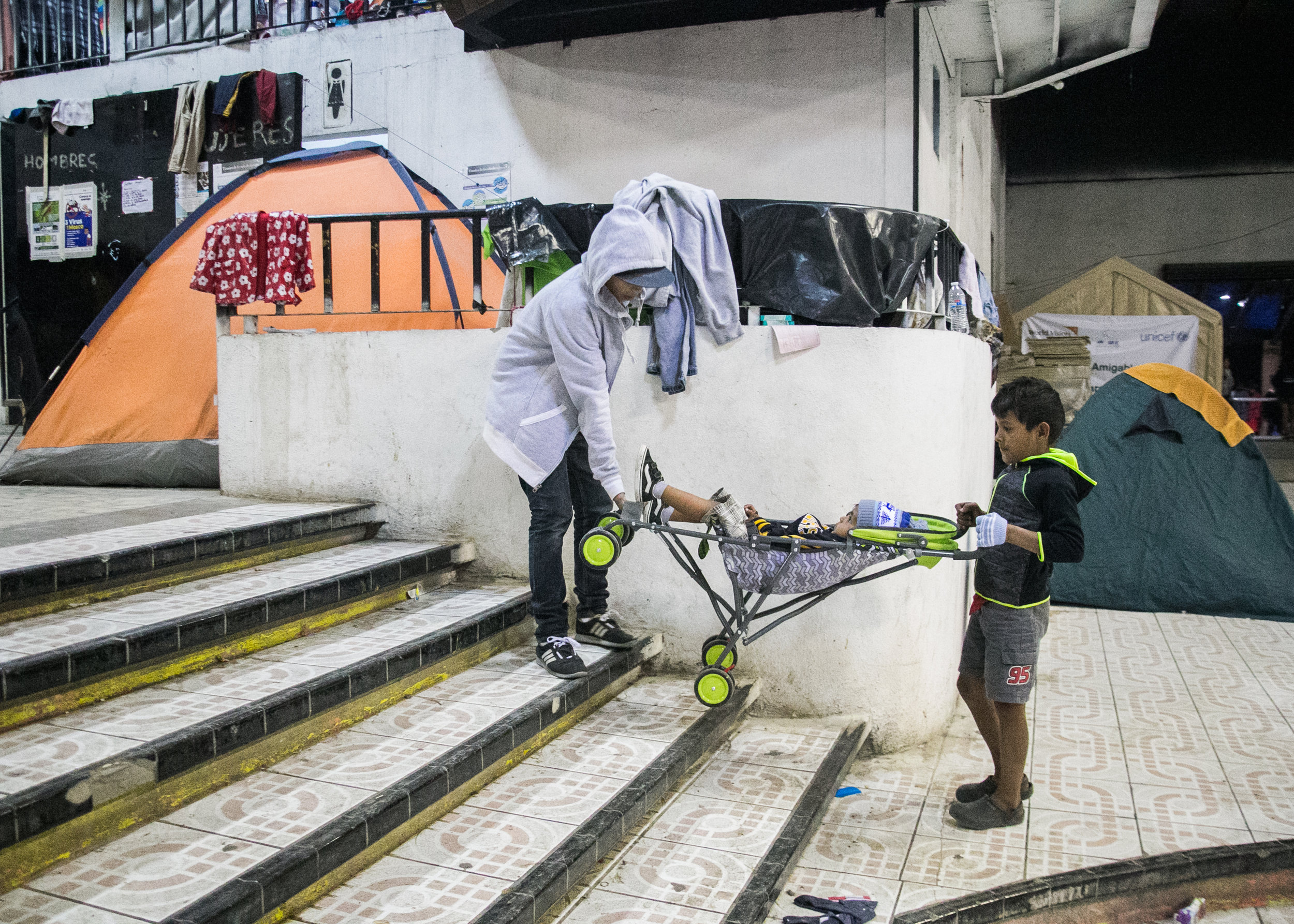  Two young boys lift their sibling’s baby stroller up a set of stairs in the Barretal refugee camp. The camp is not handicapped accessible. Renovations were not possible due to the last-second nature of the migrant caravan relocation. Over 3,000 refu