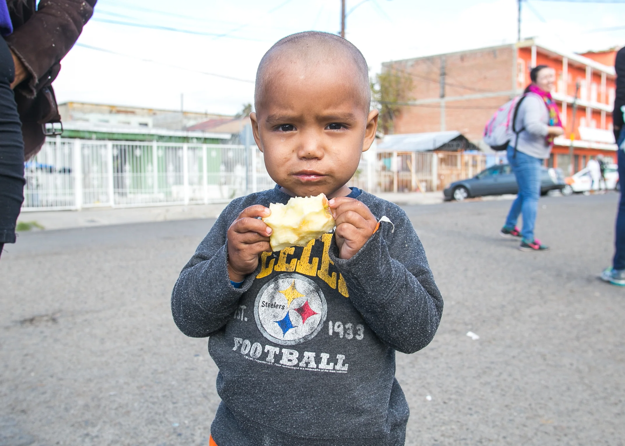 A young Honduran refugee sporting a Steelers sweatshirt eats an apple provided by World Central Kitchen at the El Chinchetta refugee compound in Central Tijuana.