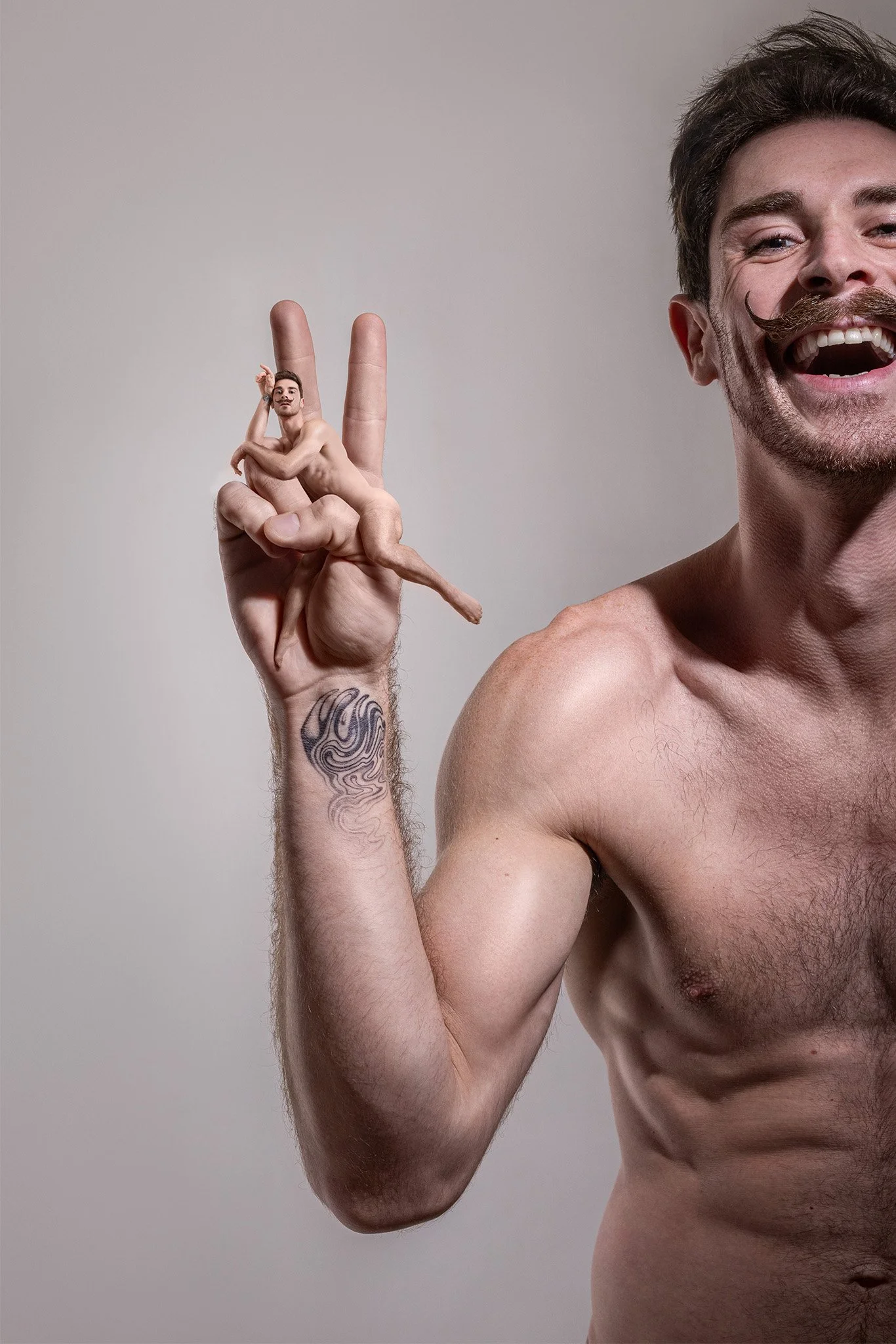Un homme nu avec une moustache, souriant, et un tatouage sur l'avant-bras, tenant une petite personne nue sur ses doigts en forme de geste peace.