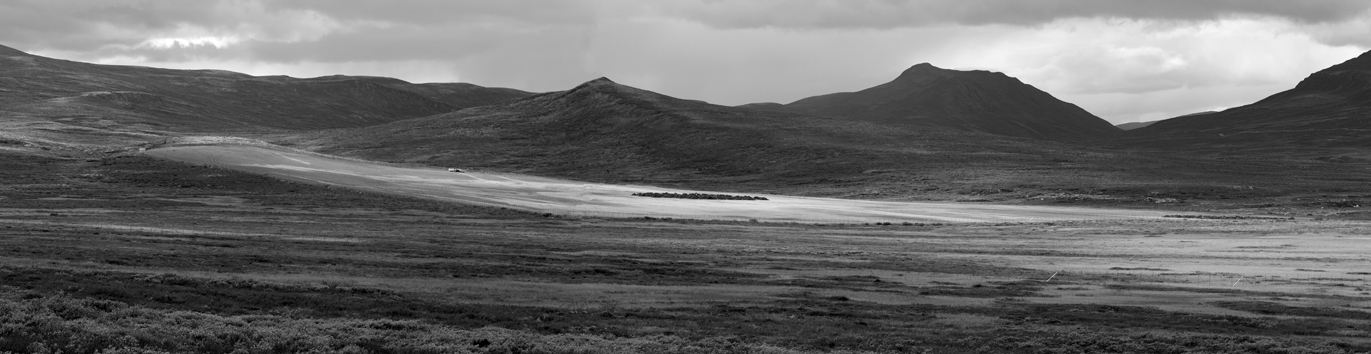 The large surface is enormous, even in this wide landscape, with perfect constructed outlines. It is an absolute rectangle, covered by gravel. The surrounding mountains leap gently down towards the valley where the square is situated, and they do not seem to belong to the same image. The square looks like a landing site for spaceships from alien galaxies. There is a small gravel road leading to the perimeter of the square, and there are traces of what must be vehicles scattered over it. In the middle you can see something that could be a small village made out of mud huts, but at closer range it is obvious that they are mounds of soil, each exactly the same size. On some of them green grass has started to grow. Apart from that, the square is desolate, without any vegetation. 