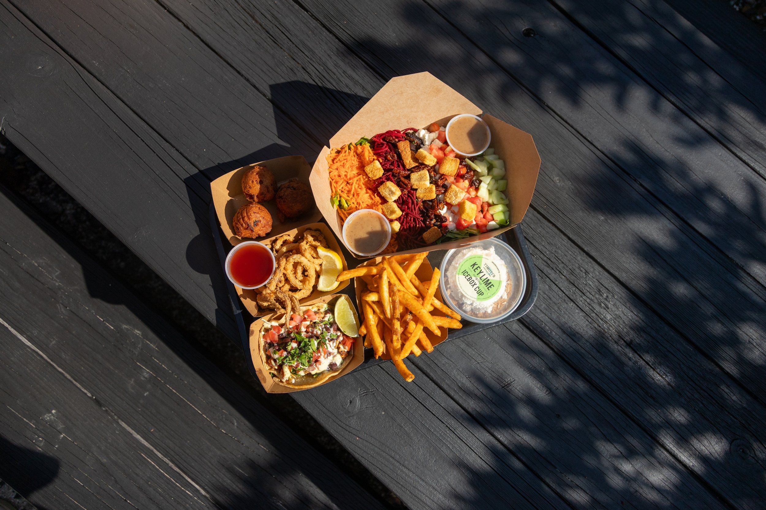 An overhead view of Timoti's food including a chopped salad box, key lime cup, hush puppies, french fries, and tacos.