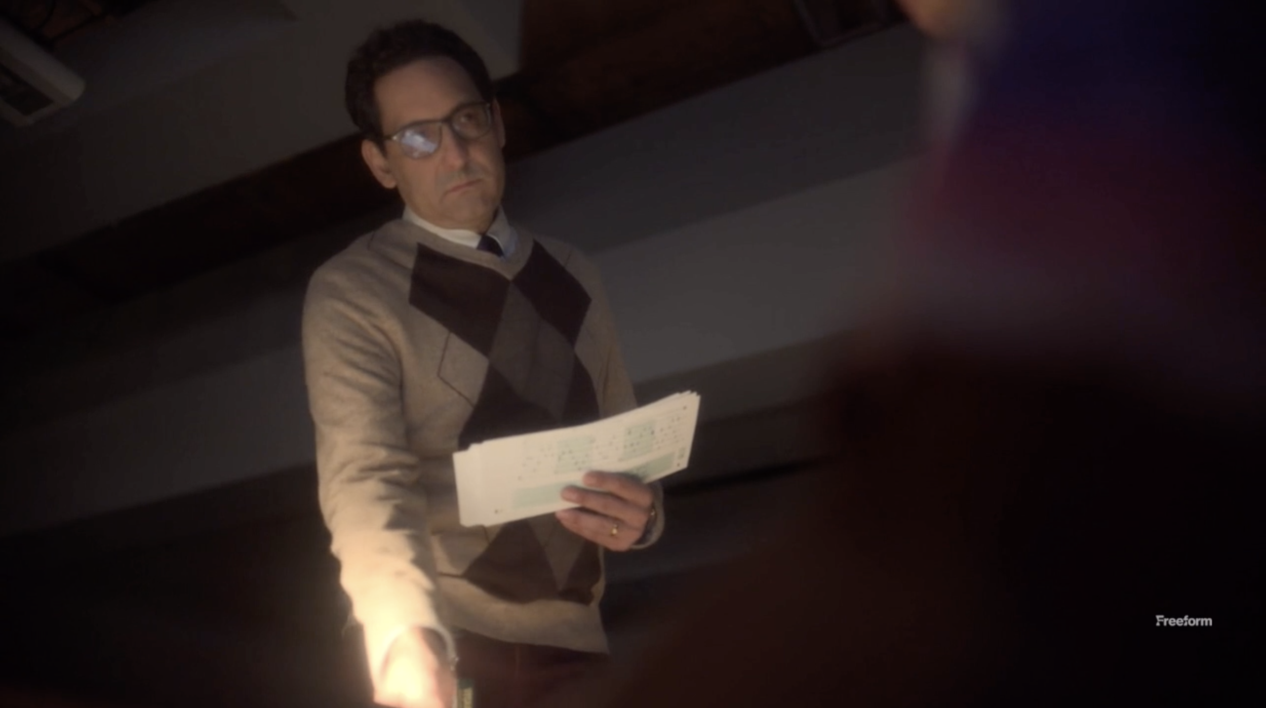 Man in glasses wearing an argyle sweater holding papers, standing indoors.