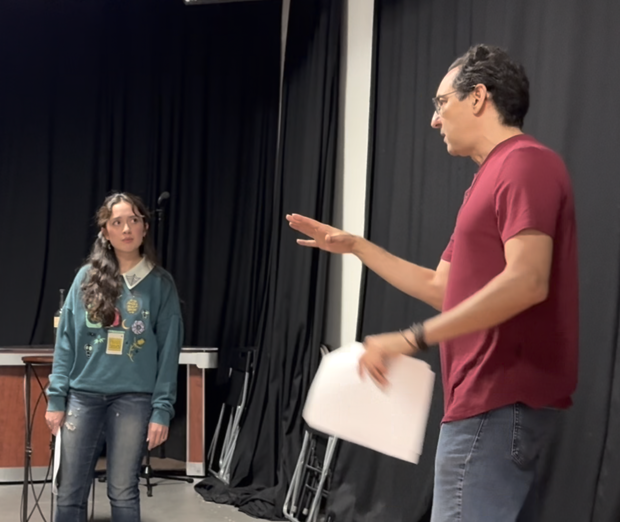 Two people in conversation, with one holding papers, in a room with black curtains and chairs in the background.