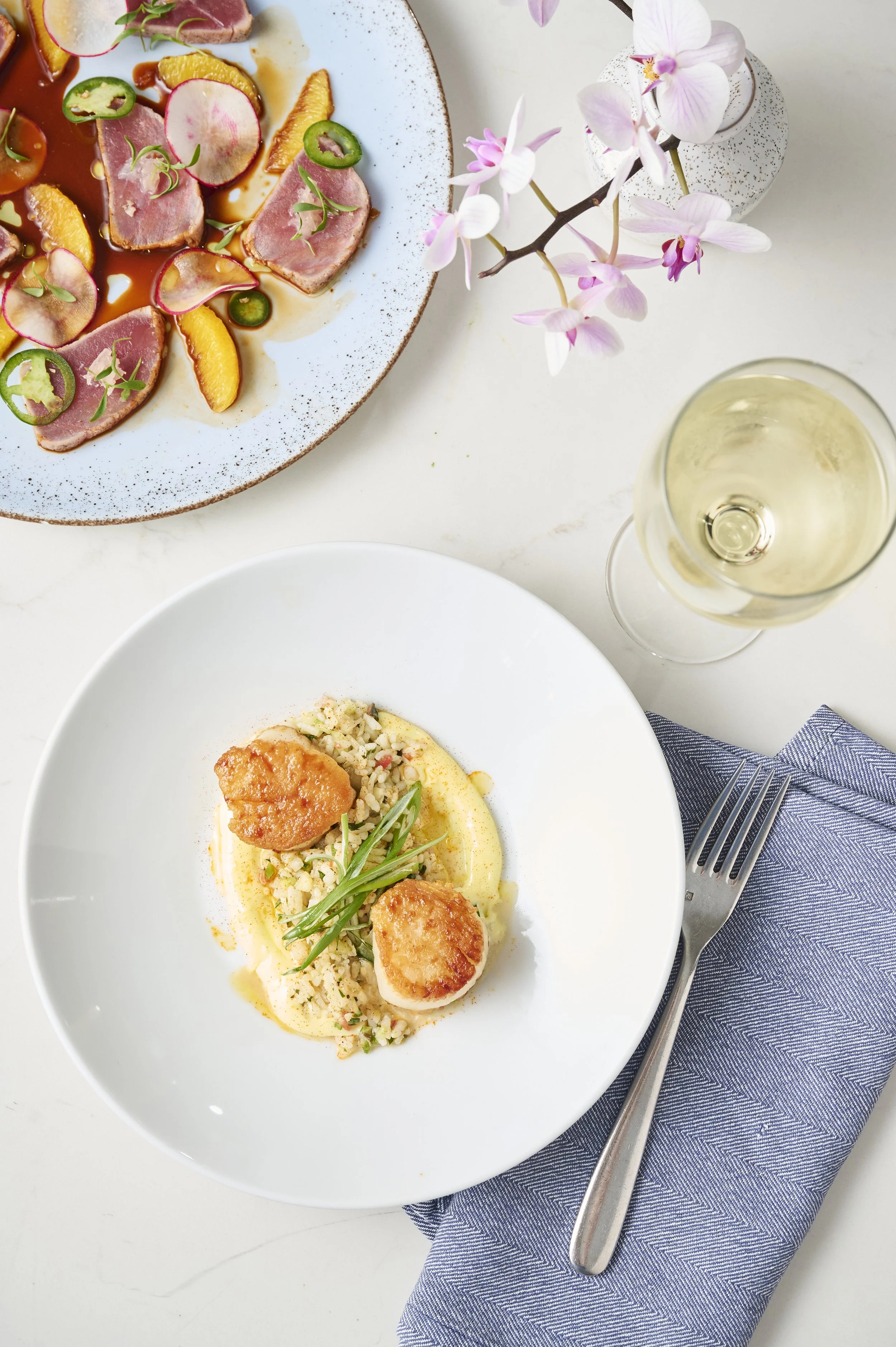 A white plate with a plated dish featuring scallops topped with green garnish, served on a bed of rice and sauce, with a sprig of green herbs. Next to the plate is a fork on a blue cloth napkin, and in the background, there is a partial view of another dish, a wine glass filled with white wine, and a vase with pink flowers on a white surface.