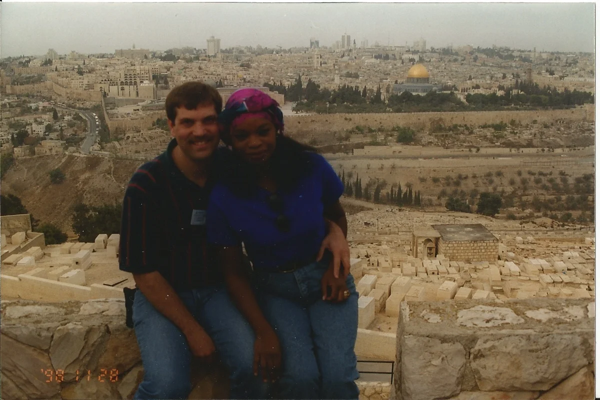 A man and a woman are sitting close together on a stone ledge overlooking a city with historical buildings and a gold dome, in a desert-like landscape with scattered trees and roads.
