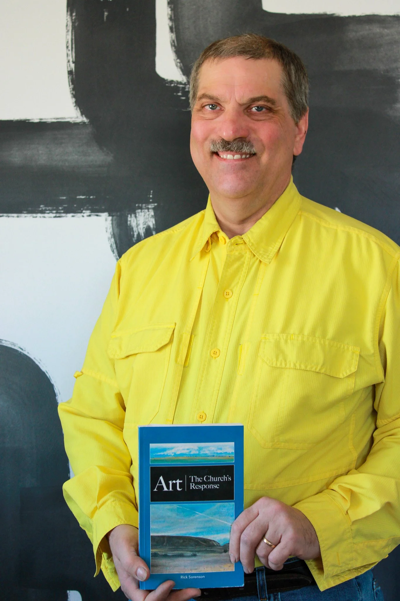 A man in a bright yellow shirt holding a book titled "Art: The Church's Response" by Rick Sorenson, standing in front of a dark background with abstract white and black paint strokes.
