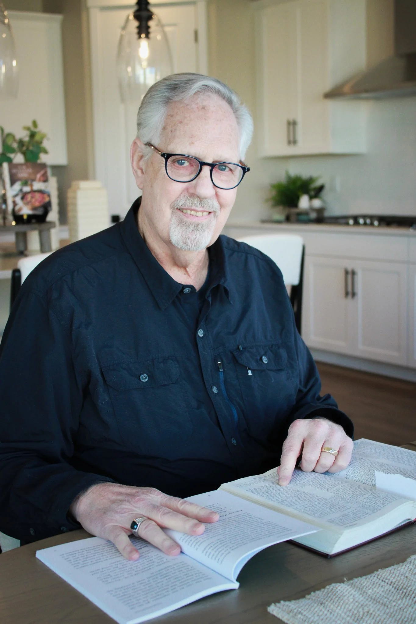 An elderly man with gray hair, glasses, and a goatee is sitting at a table in a kitchen while reading a large book. He is pointing to a specific line or passage in the book with his right hand. The kitchen background includes white cabinets, a potted plant, and some decorative items.