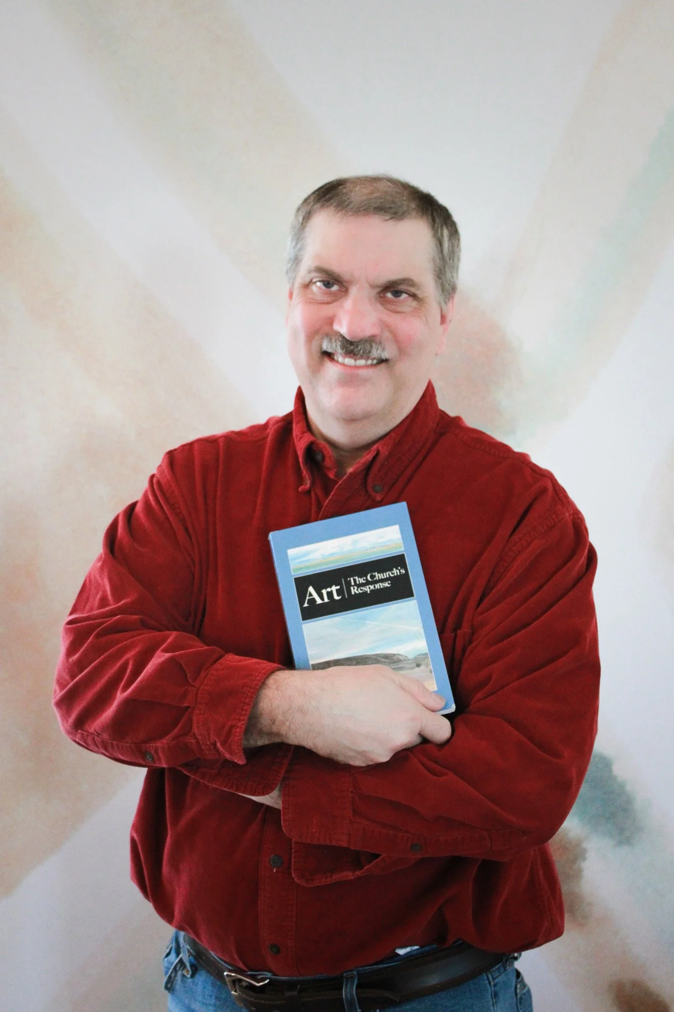 A man with a mustache and short hair wearing a red jacket, holding a blue book titled 'Art: The Church's Response,' standing in front of a plain background.