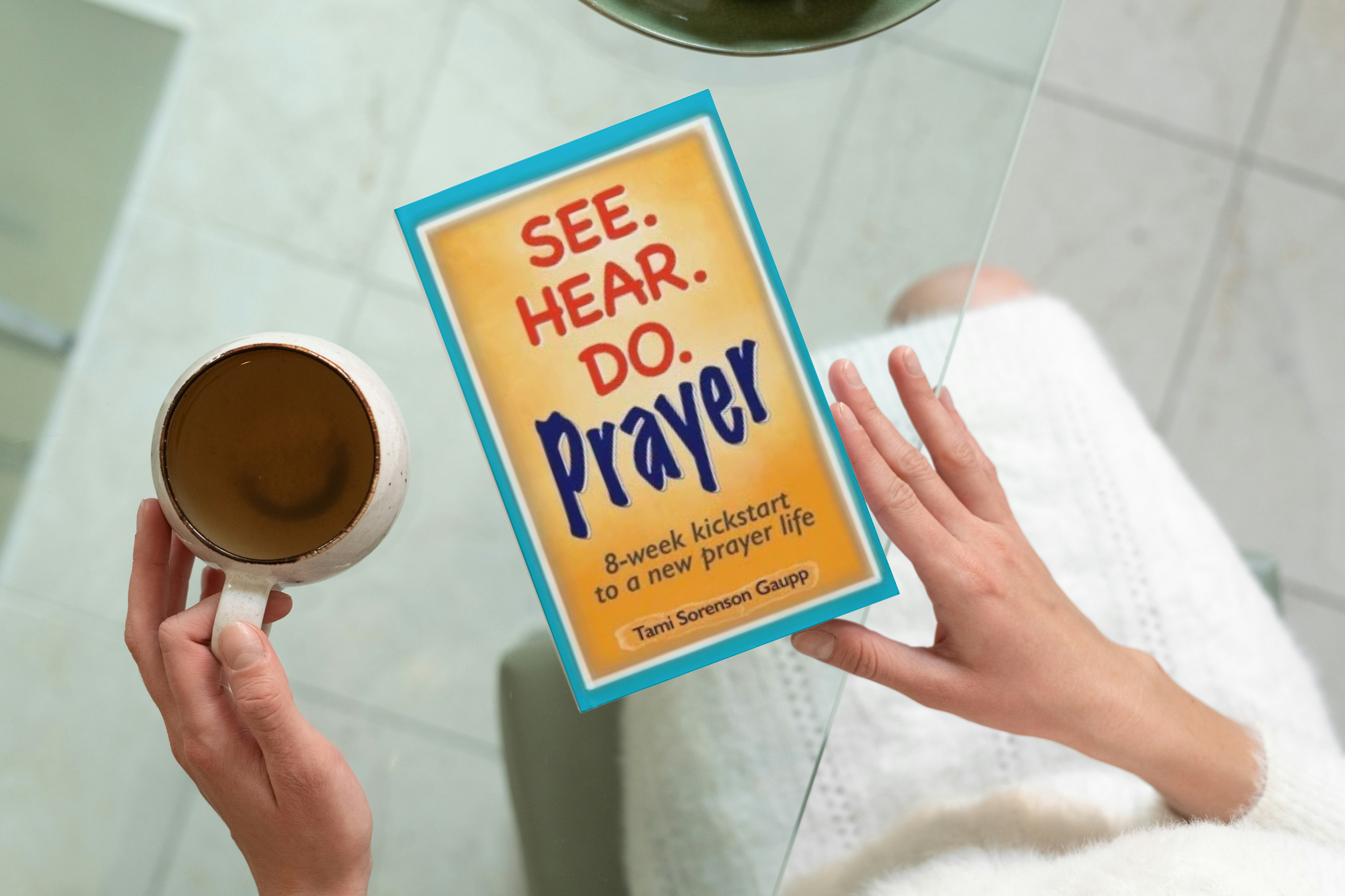 The image shows two hands holding a cup of coffee and a book titled "See. Hear. Do. Prayer:" by Tami Sorenson Gaup, on a glass table.