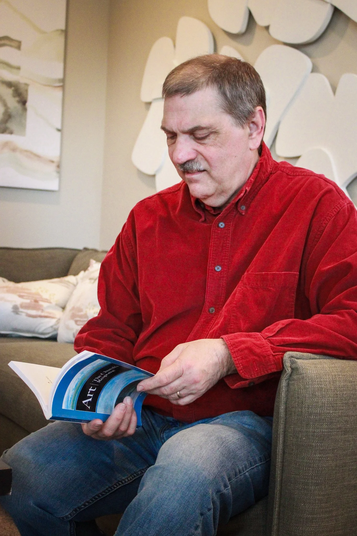 A middle-aged man with short brown hair and a mustache wearing a red long-sleeve shirt, sitting on a beige sofa, reading a brochure or magazine. The background features wall art with abstract white shapes and a small painting. There is a plush pillow beside him.