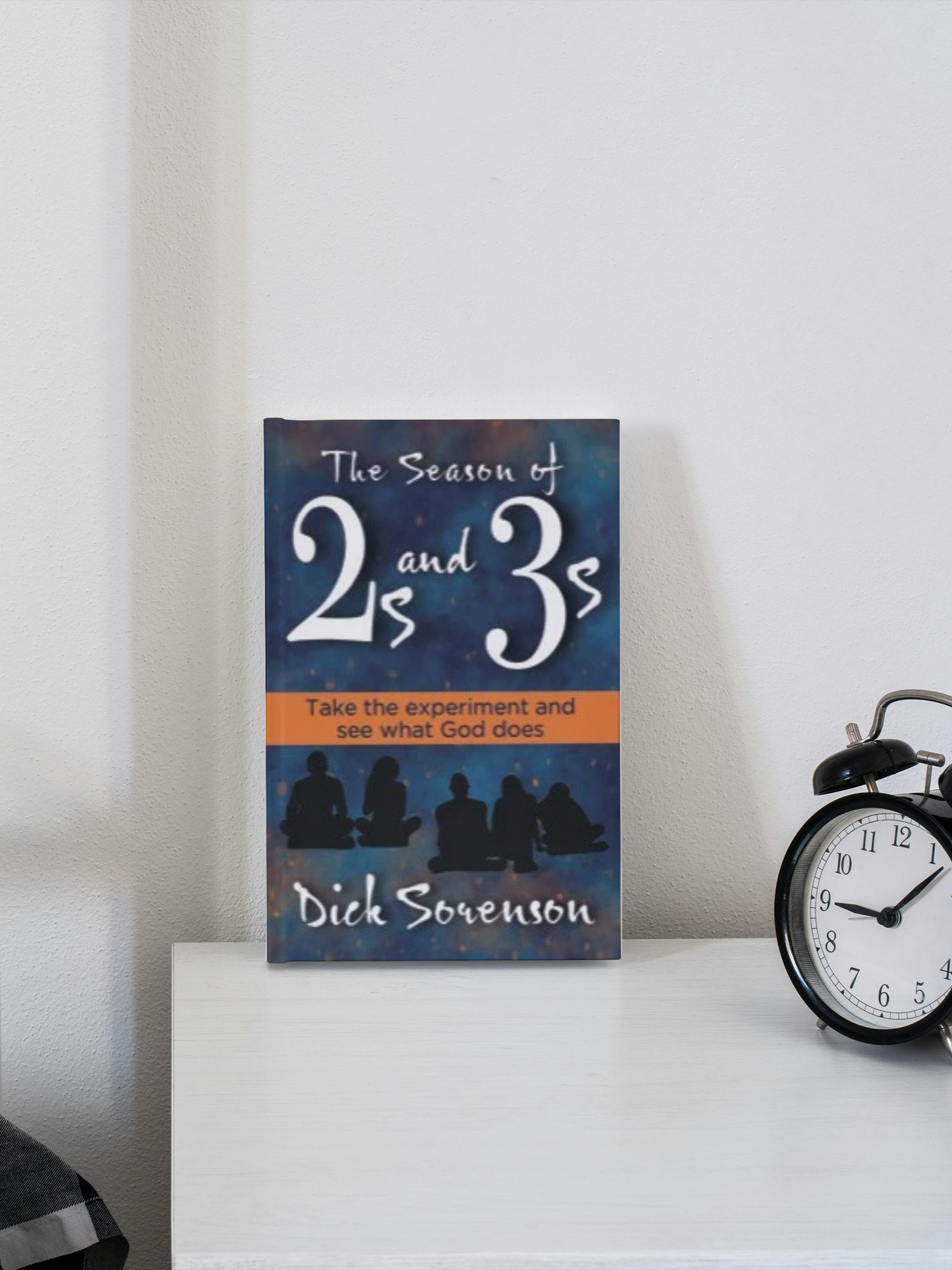 A book titled 'The Season of 2s and 3s' by Dick Souenson is placed on a white table next to a black and white alarm clock. The book cover is blue with silhouettes of people sitting cross-legged, and an orange band with black text.