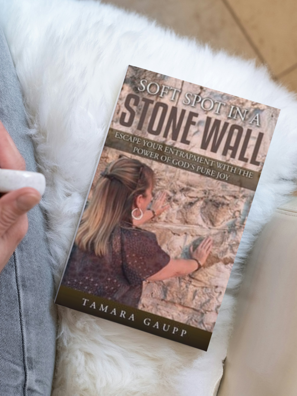 A woman with long hair looking at a stone wall with her hands raised and touching it. The image is part of a book cover titled 'Soft Spot in a Stone Wall' by Tamara Gaup