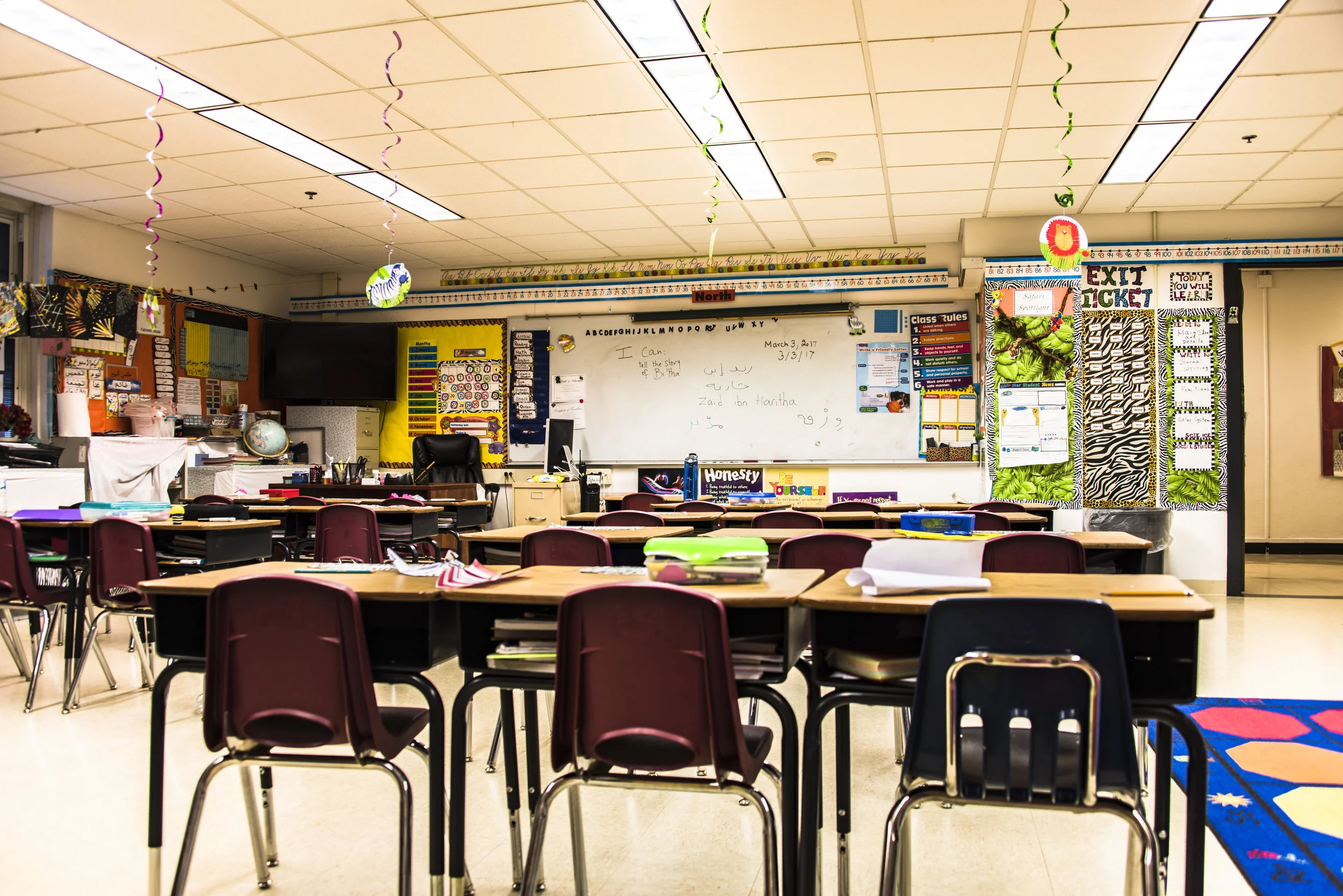 Image: An empty elementary school classroom with desks, a white board, and posters on the walls. 