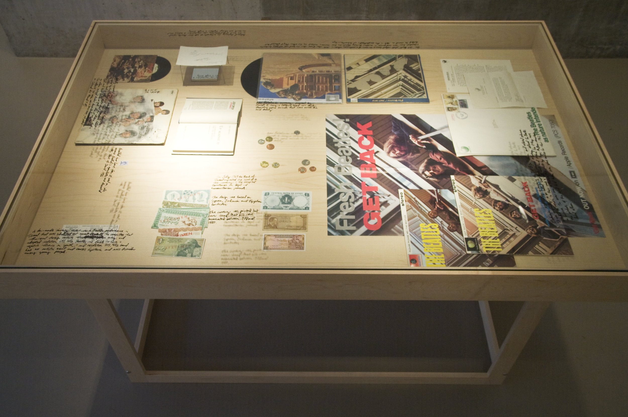   Fastasy items, from unreleased Beatles albums to proposed Palestinian currency are presented in a vitrine with handwritten captions on glass.    