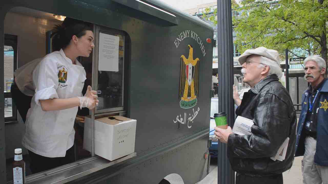 Enemy Kitchen is now a fully functional food truck on the streets of Chicago that features Iraqi refugee cooks and US veterans of the Iraq War serving as sous chefs. In this way, the power dynamic in Iraq is inverted, as Americans now take orders from the Iraqis. The truck features the Chicago flag, rendered in Iraqi colors and serves food on paper replicas of the plates looted from Saddam Hussein's palaces in 2003. (add to new image - slide 93) The truck has made visible and audible the people on both sides of the war, as dialogue accompanies the serving and consumption of food, and the truck creates a critical social space. 