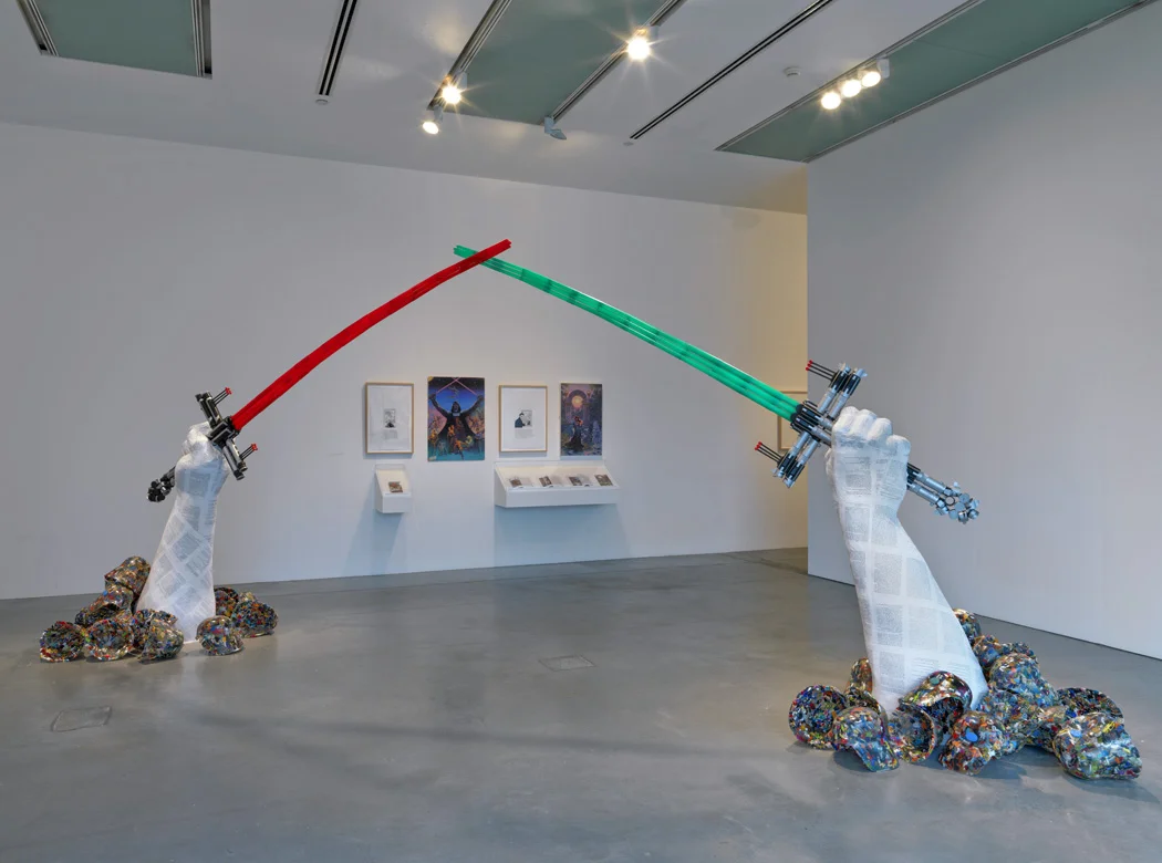 The installation’s title derives from a saying of Hussein’s that appeared on the invitation card for the inauguration of the Arch, built in 1989 to commemorate Iraq’s “victory” over Iran. The original monument features swords cast from weapons used in the Iran-Iraq War and features 2,500 helmets of vanquished Iranians cascading at its base. A large-scale sculpture reproduces this monument in materials that connect with the theme of boyhood fantasy: dozens of plastic light sabers for the Arch, GI Joe figurines encased in resin for the helmets (modeled on the Fedayeen’s own Vader-like helmets) that surround the sculpture’s base.