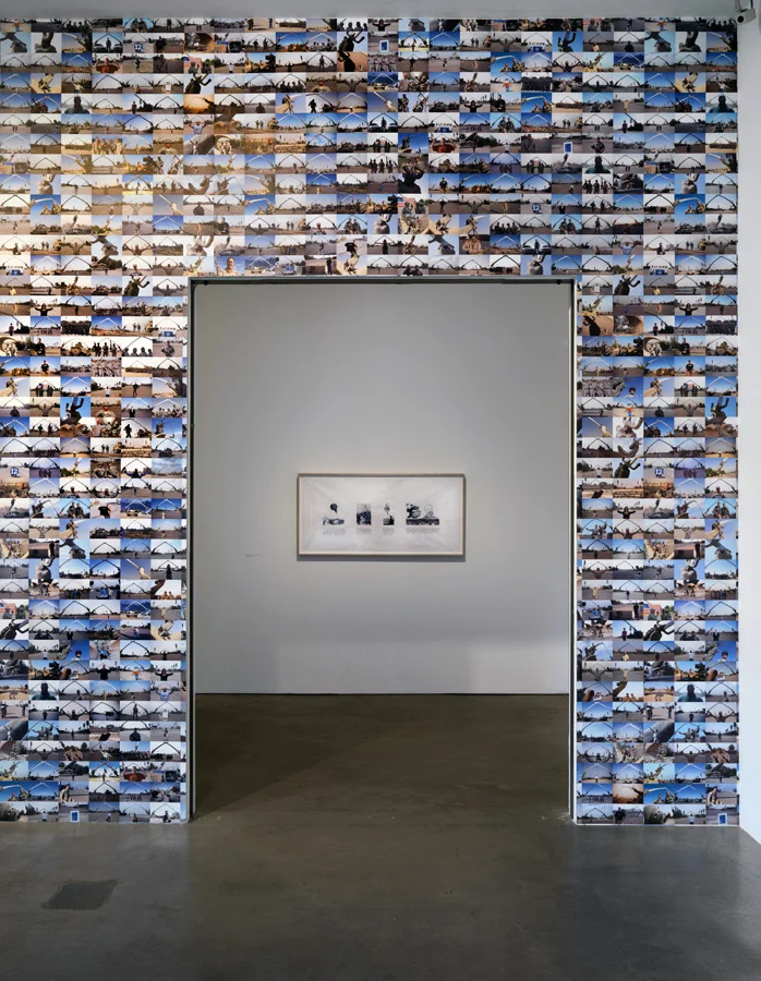 The entrance to the exhibition space is framed by wallpaper featuring the hundreds of photographs found on soldiers’ blogs, Flickr accounts and other sources that feature military officers and contractors posing in front of the Victory Arch.