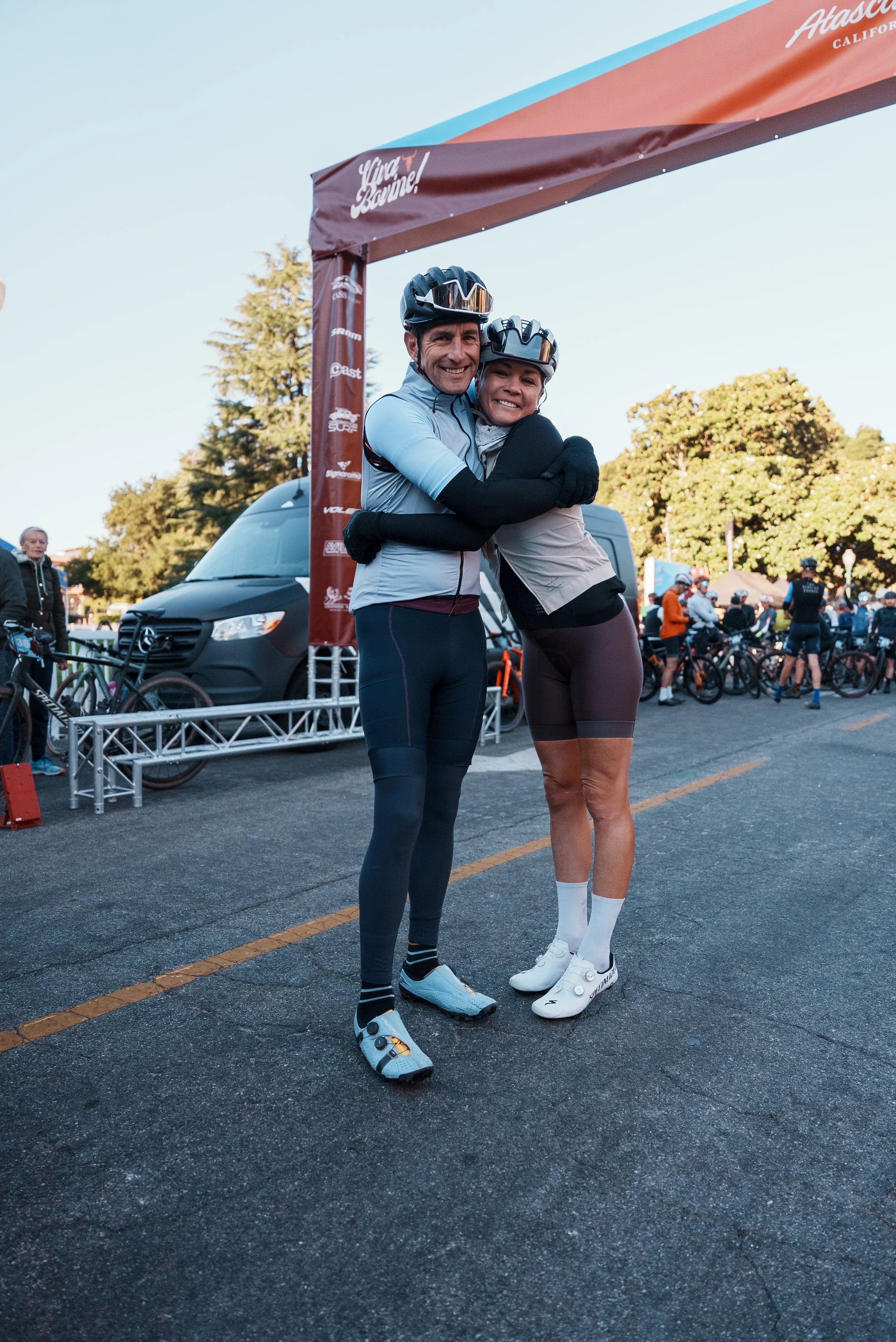 Two cyclists hugging under a starting arch at an outdoor cycling event, wearing helmets and cycling gear, with other cyclists and a van in the background.