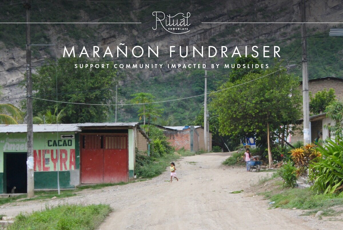 Bright red &amp; green brick buildings divided by a dirt road, nestled against the backdrop of mountains. A child runs across the road while one is using a wheelbarrow. The caption reads Marañon Fundraiser—Support Community Impacted By Mudslide.