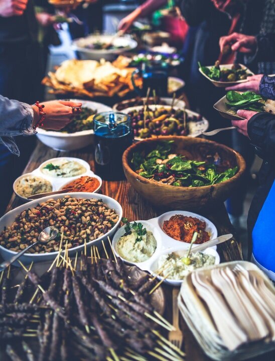 A decadent dinner party table, with a wide variety of foods. Closest in the foreground are skewers, a bean salad, greens, and various roasted vegetable dips. In the background out of focus people are serving food.