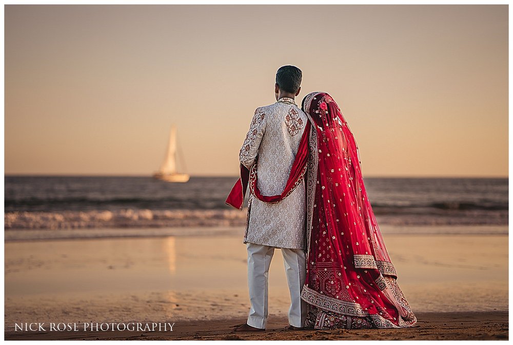  A stunning sunset portrait of the bride and groom, captured by destination Indian wedding photography, with the ocean and sky painted in vibrant shades of orange and pink. 