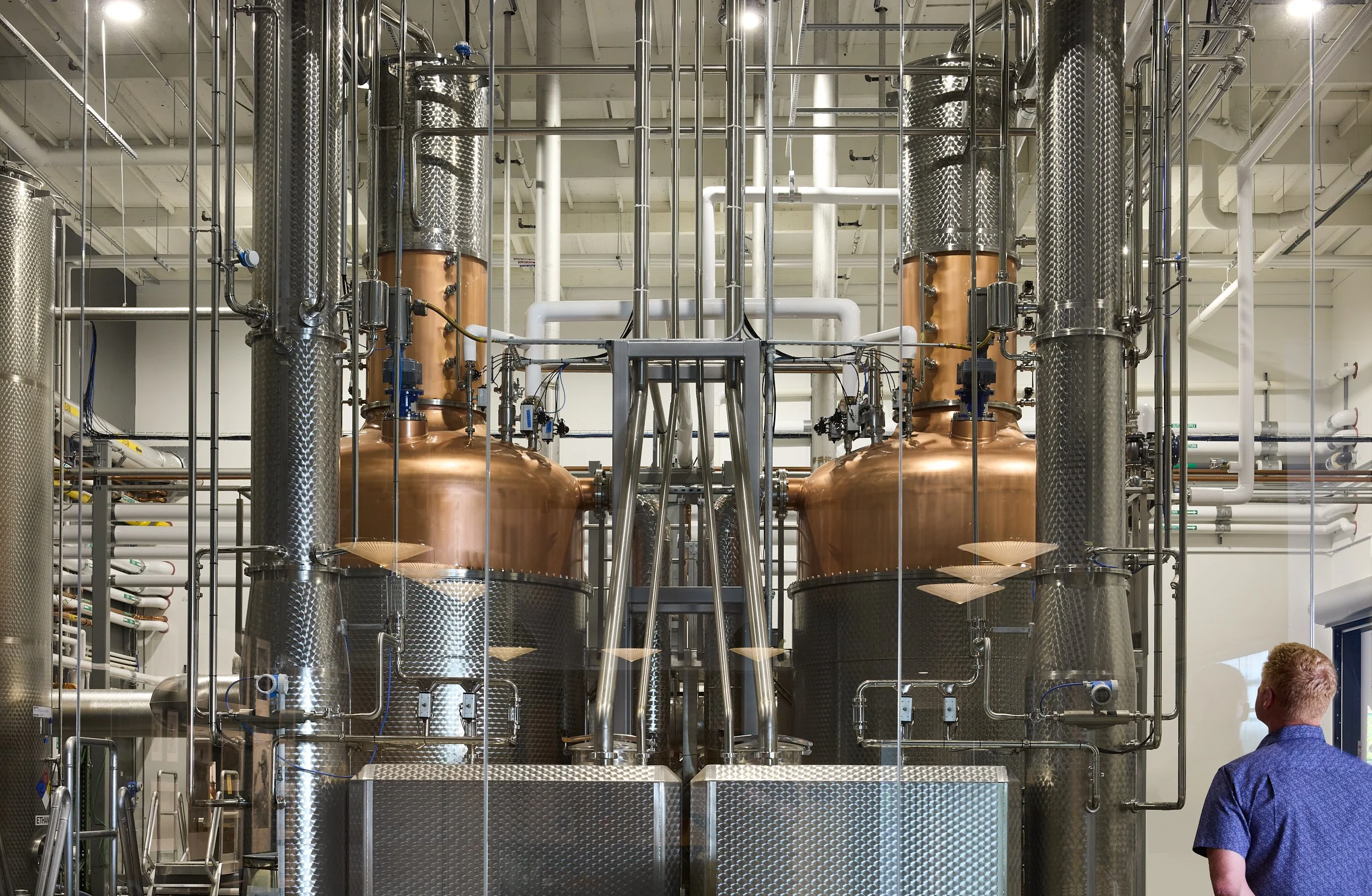 Interior architectural image of visitor looking through glass wall at Aviation Gin distillery production equipment in Portland, Oregon, designed by fi architecture.