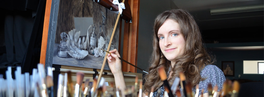 A woman sitting at an easel painting with brushes in the foreground.