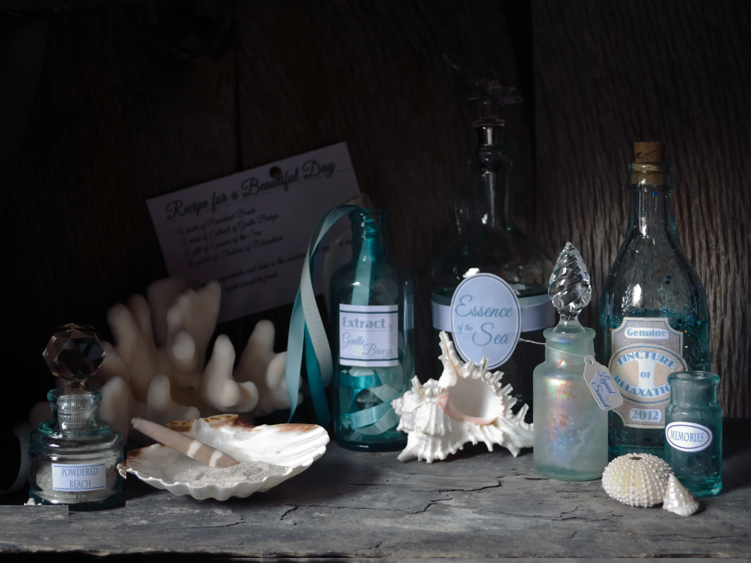 Because this was a story about the sea, I needed a couple more sea creatures, and therefore, I added the small white and pink Murex Ramosus shell. I like to repeat textures, shapes, and colors throughout my compositions in order to both lead the eye and create more visual harmony. The coral was a good match for repeating the spikes of the Murex shell as well as being similar in color and texture to the sea urchin. The pink in the Murex shell also complemented the pinks in the bottle labeled "Liquid Sun Set" as well as the subtle pink in the sea urchin carapace. The brown in the shell I used to symbolize a mixing bowl harmonized nicely with the cork that I had tinted to match in a bottle across from it in the composition. The brown sea urchin spike in the same shell was also useful for symbolizing a mixing spoon. I replaced the small white and brown murex shell for one that was more conical to lead the eye back into the composition and also repeat the similar shape of one of the glass stoppers. Finally, I moved the bottle labeled powdered beach to the left side of the composition to balance the bottles in both color and texture. I then adjusted my black velvet curtain over part of the front of my shadow box so that it created a more interesting lighting effect on the objects. I made sure that both the visually busy recipe card and highly textured weathered wood panels were in more shadow and would recede into the background making the foreground objects more pronounced. At this point I felt satisfied with my still life composition and was ready to move onto the next step...