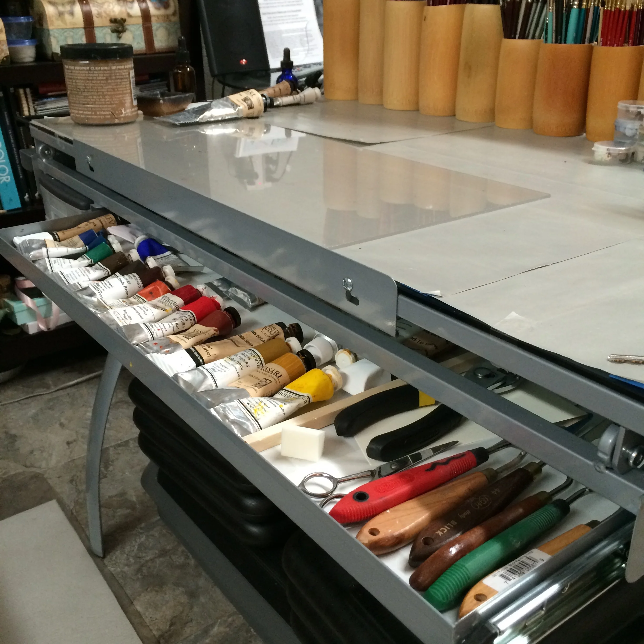 A desk drawer full of paint tubes and tools and brushes on the table top.