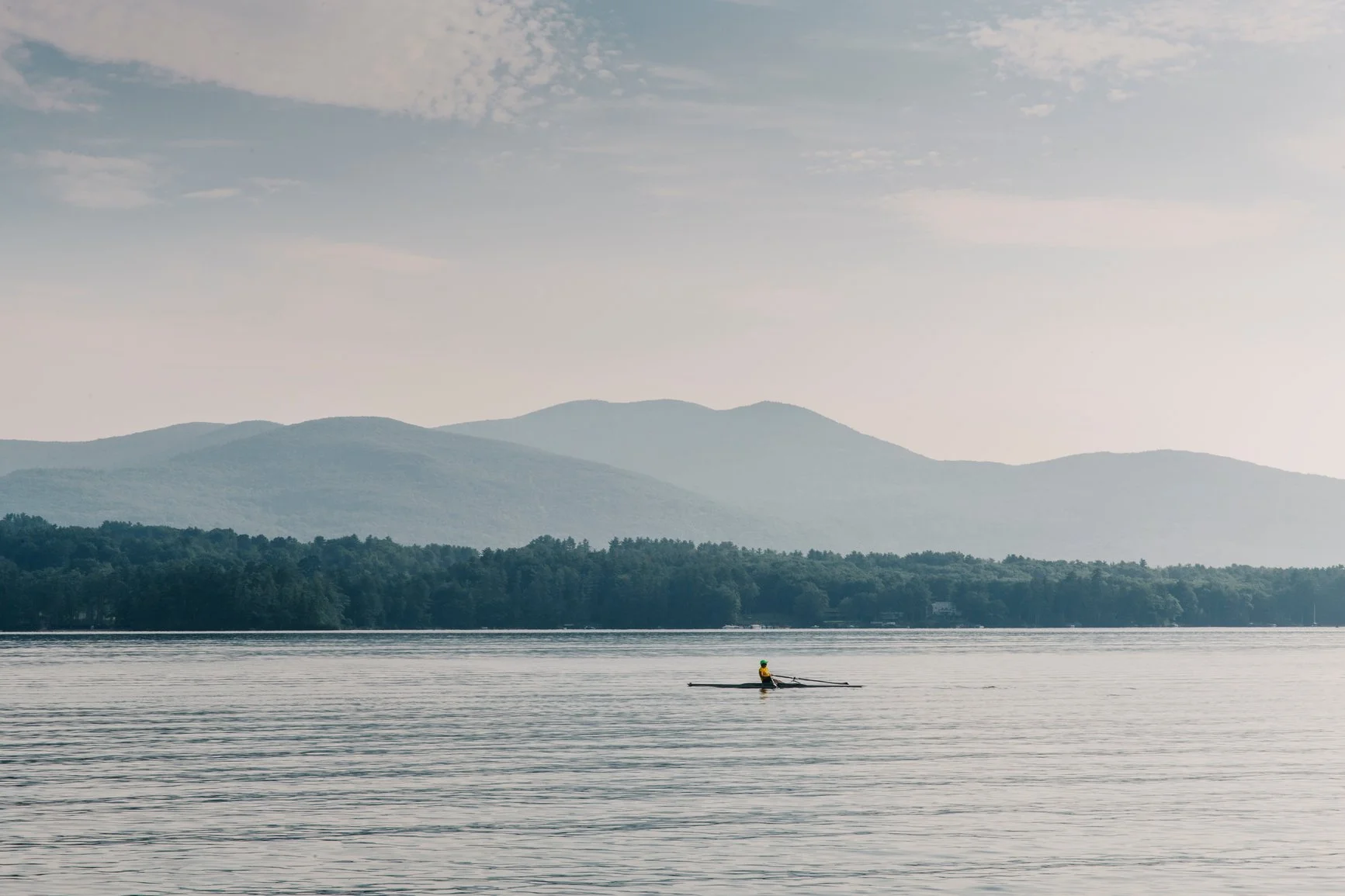 A person kayaking on a calm lake with mountains in the background and a partly cloudy sky.