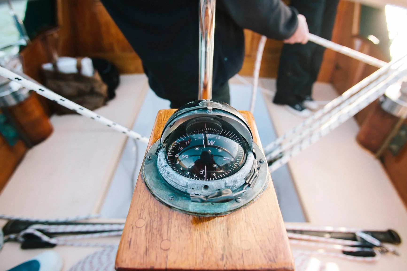 A vintage nautical compass mounted on a wooden platform, with a person's legs and a ladder visible in the background.