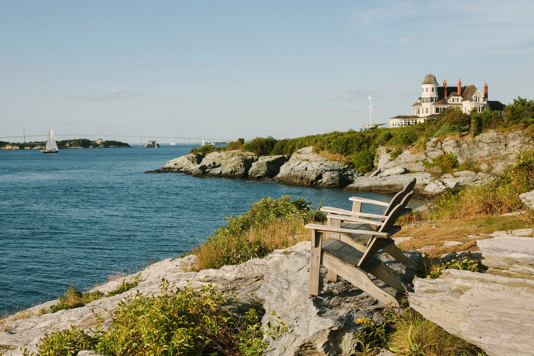 View of a rocky coastline with two wooden Adirondack chairs facing the water, a house on the hill, sailboats on the water, and a bridge in the distance.