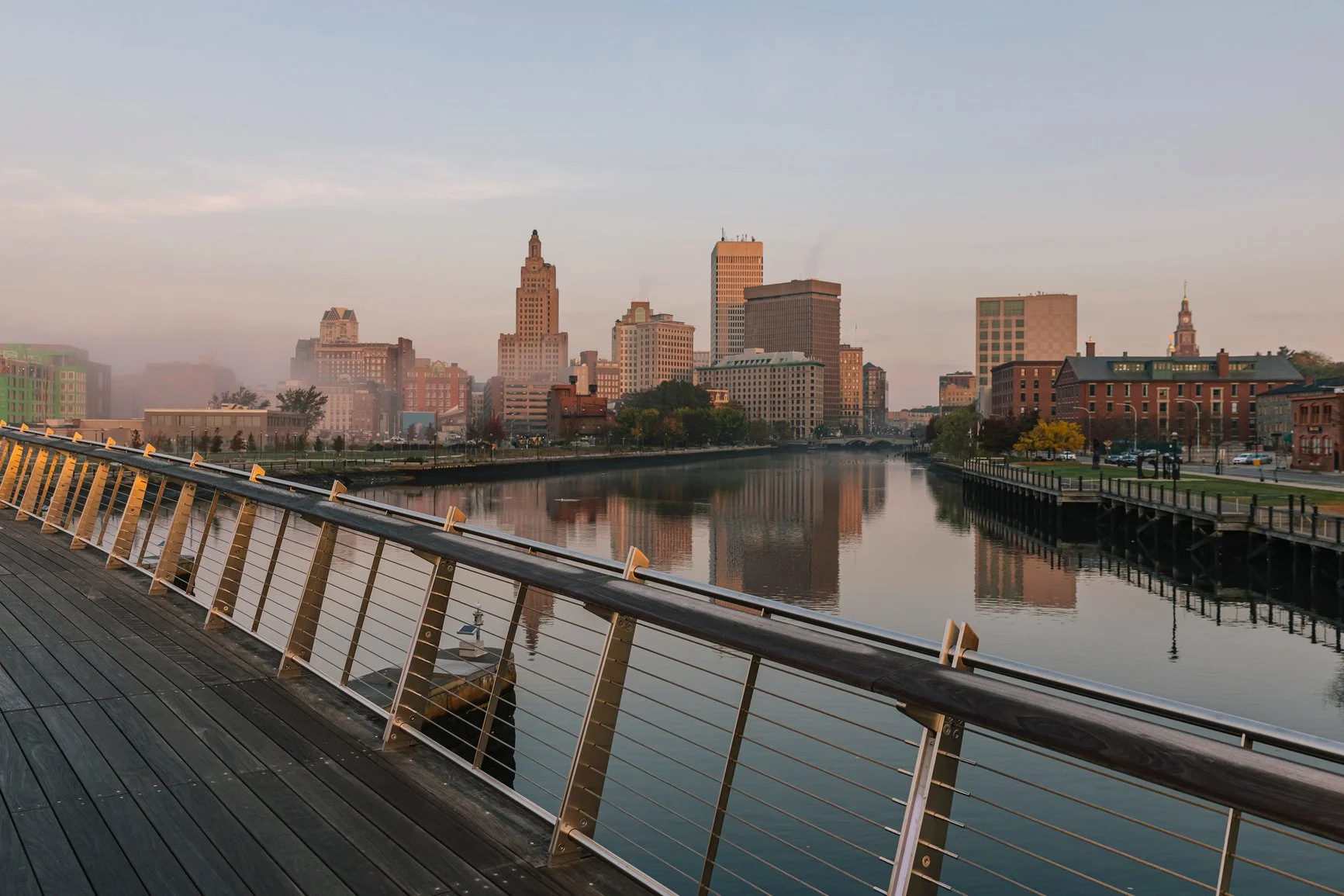 City skyline with tall buildings reflected in a river, taken from a wooden boardwalk with a metal railing.