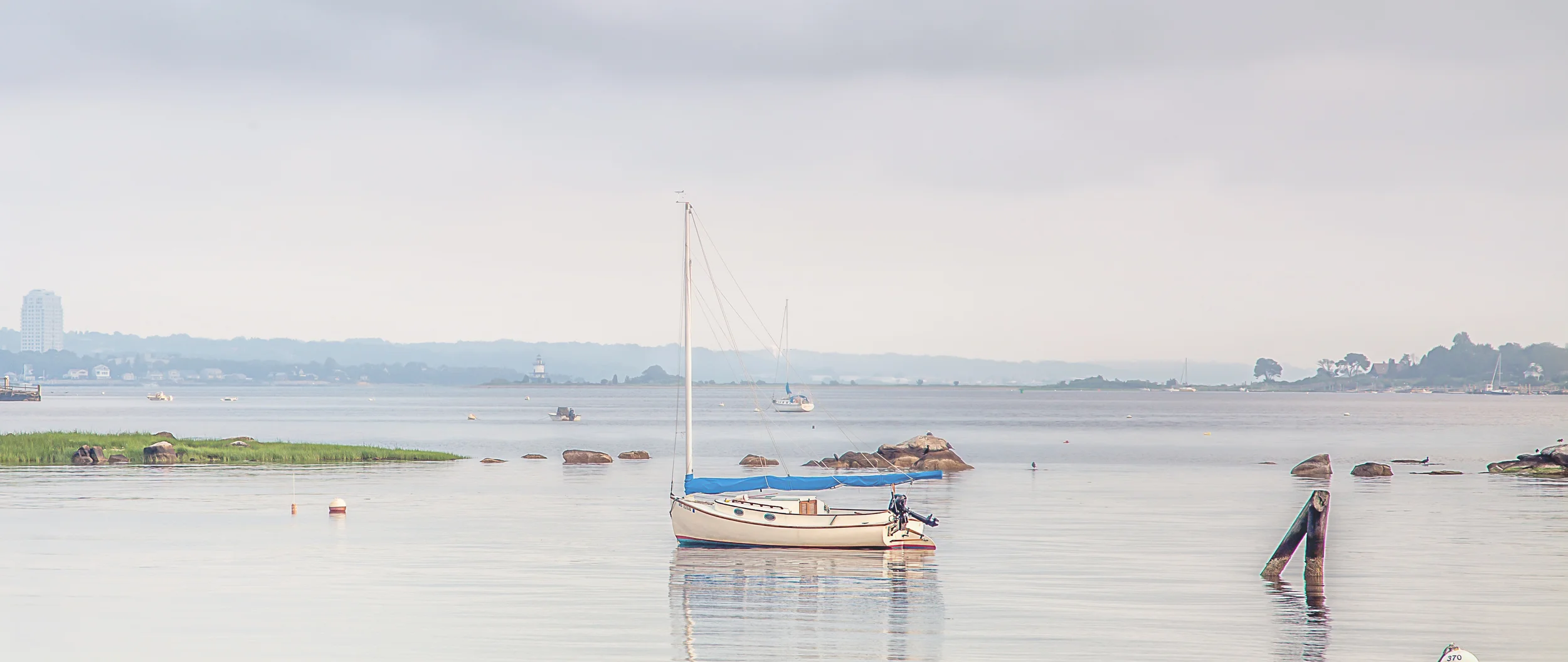 A small sailboat with a blue cover on calm water, with rocks and buoys in the foreground, and other boats, trees, and a distant cityscape in the background.