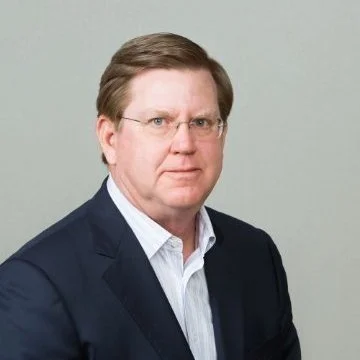 A middle-aged man with light brown hair and glasses, wearing a dark blazer and light-colored shirt, standing against a plain light background.