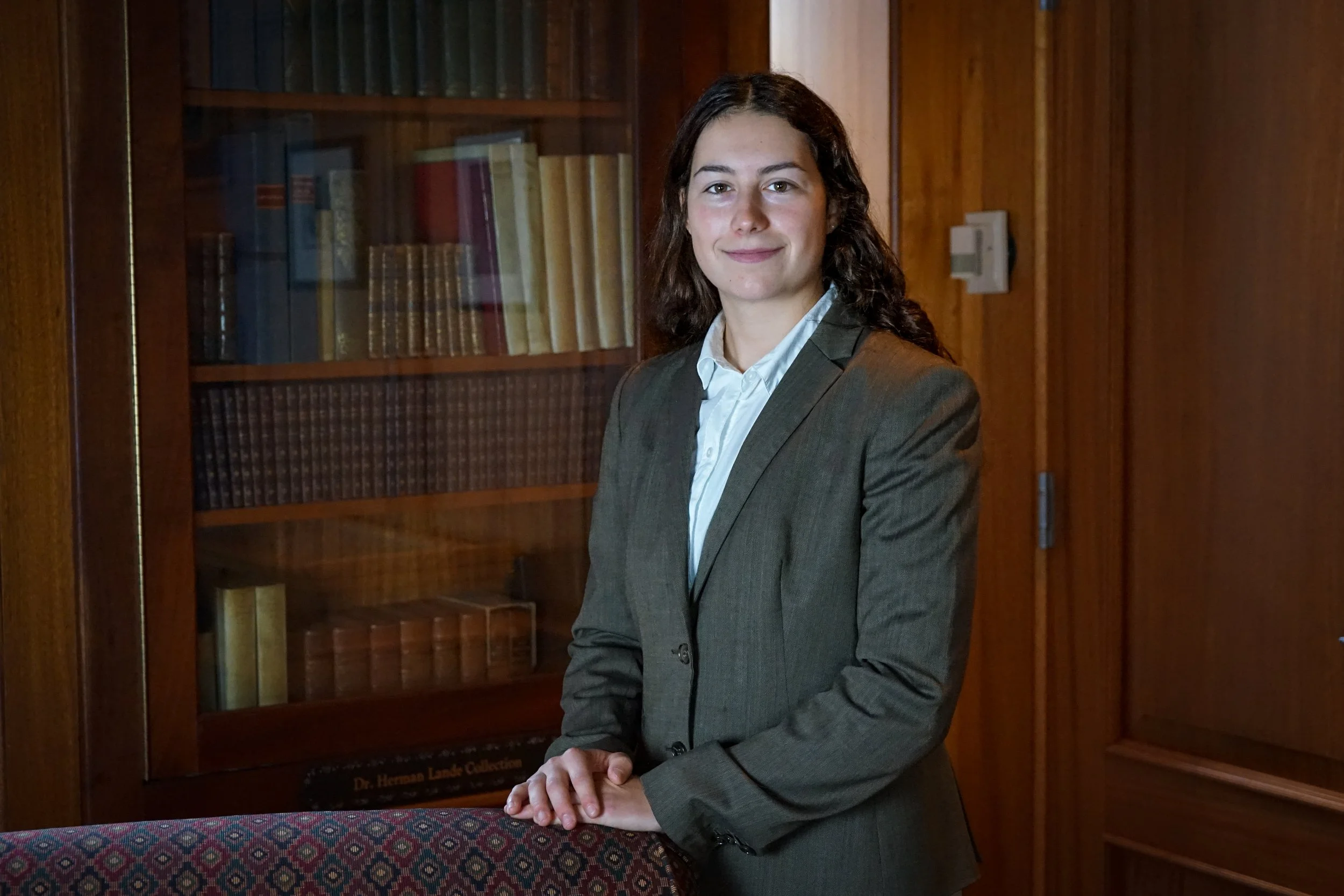 A woman with dark brown hair wearing a gray blazer and white shirt, standing in front of a wooden bookshelf filled with books, with her hands resting on a table with a colorful patterned tablecloth.