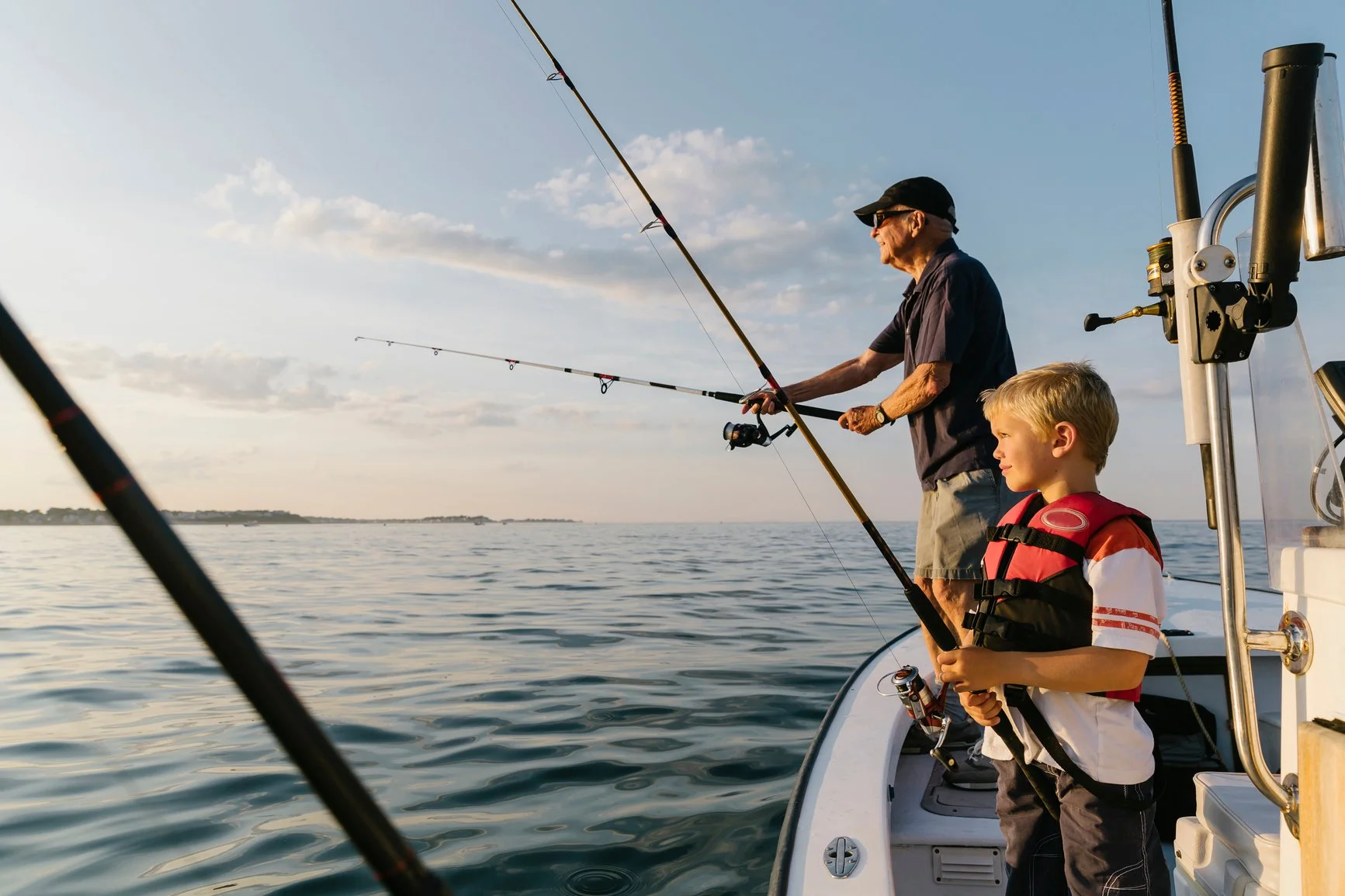 An elderly man and a young boy fishing from a boat on calm water during sunset. The elderly man is wearing a dark shirt, cap, and sunglasses, while the boy is in a red life jacket and striped shirt.