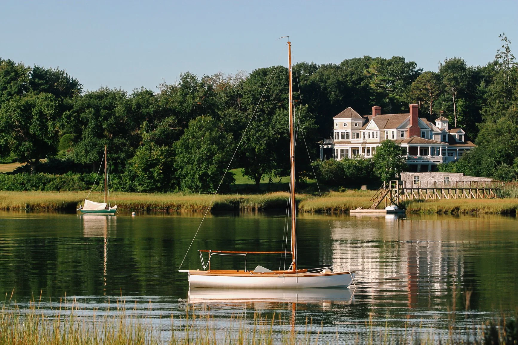 A house on a hill next to the water with a sailboat floating on the river in the foreground, surrounded by green trees.